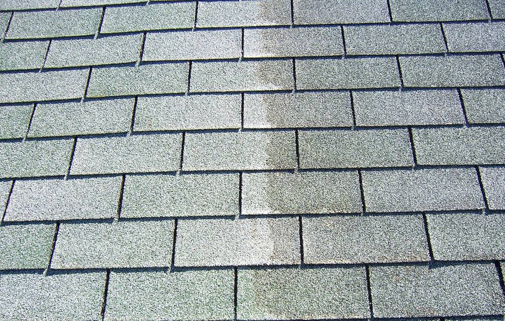 Close-up of asphalt shingles with one cleaned stripe running through the algae growth, showing the color difference